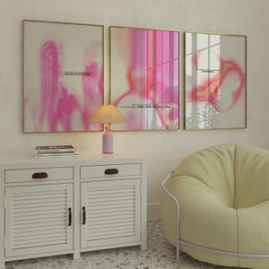 May include: Three framed abstract art prints with pink and white color gradients. The prints are displayed above a white cabinet with a lamp and a stack of books. A yellow beanbag chair is in the foreground.