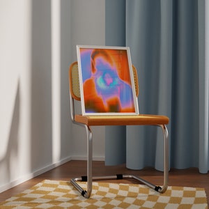 May include: A white framed print of an abstract portrait in shades of orange, blue, and purple, sitting on a wooden chair with a woven back.