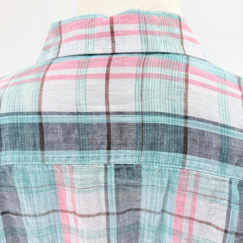 May include: Close-up of a plaid shirt collar and back yoke. The fabric features a pattern of intersecting lines in shades of pink, teal, gray, and brown on a white background. The shirt is made of a lightweight material.