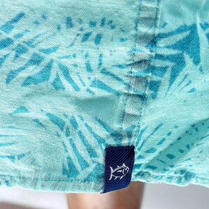 May include: A light turquoise shirt featuring a darker blue palm leaf pattern. The garment has a vertical seam running down the center and a small, rectangular navy blue fabric tag at the bottom hem, which displays a white embroidered fish icon.
