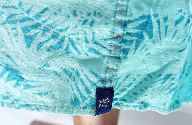 May include: A light turquoise shirt featuring a darker blue palm leaf pattern. The garment has a vertical seam running down the center and a small, rectangular navy blue fabric tag at the bottom hem, which displays a white embroidered fish icon.