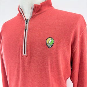 May include: A coral-colored long-sleeve pullover shirt with a high collar and a partial zipper. The shirt features a small embroidered emblem of a skull with a lightning bolt design in yellow and green.