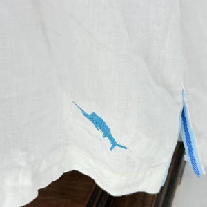 May include: White linen shirt with a blue marlin embroidery on the bottom. The shirt has a blue and white striped detail on the side. The fabric appears to be lightweight and breathable.