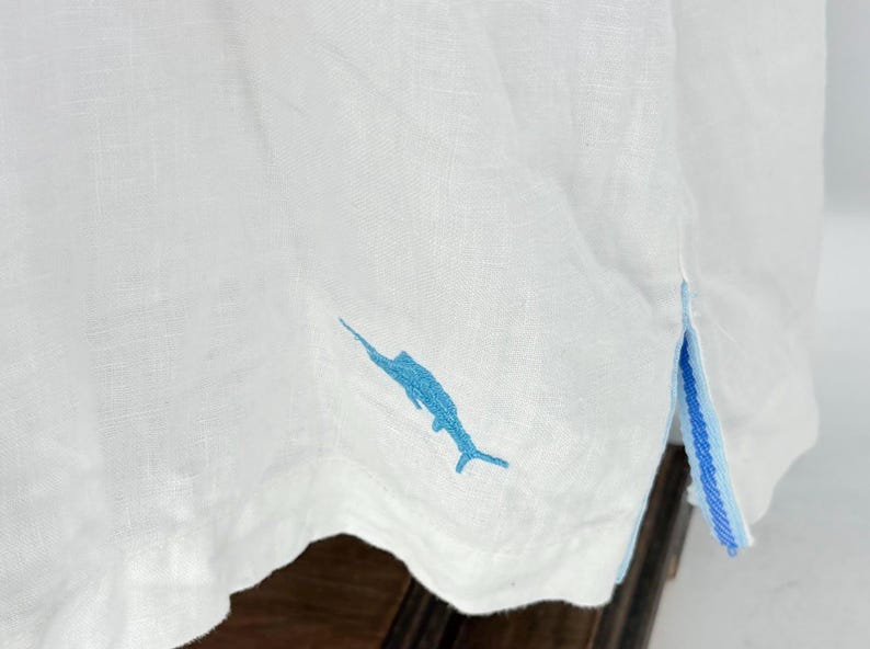 May include: White linen shirt with a blue marlin embroidery on the bottom. The shirt has a blue and white striped detail on the side. The fabric appears to be lightweight and breathable.