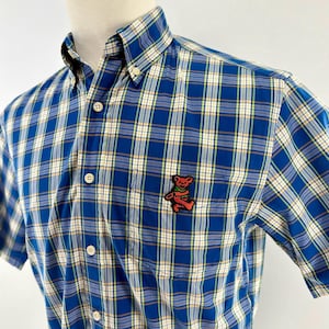 May include: Blue plaid button-down shirt with short sleeves. The shirt features a small pocket on the chest and a colorful bear patch. The plaid pattern includes white, yellow, and tan lines. The shirt is buttoned up and has a collar.