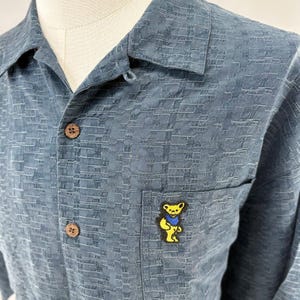 K&ouml;nnte beinhalten: Ein blaues Hemd mit Kn&ouml;pfen, einem strukturierten Muster und einer kleinen Tasche auf der Brust. Die Tasche hat eine gelbe B&auml;rennadel mit einer blauen Fliege. Das Hemd hat Kn&ouml;pfe in Holzoptik.