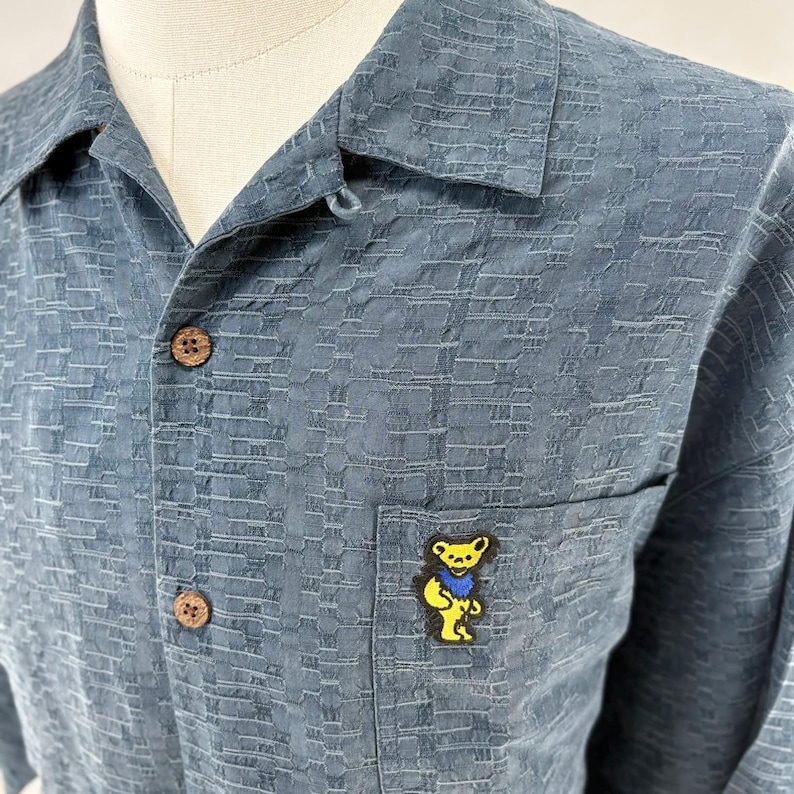 K&ouml;nnte beinhalten: Ein blaues Hemd mit Kn&ouml;pfen, einem strukturierten Muster und einer kleinen Tasche auf der Brust. Die Tasche hat eine gelbe B&auml;rennadel mit einer blauen Fliege. Das Hemd hat Kn&ouml;pfe in Holzoptik.