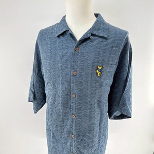 K&ouml;nnte beinhalten: Blaues kurz&auml;rmeliges Hemd mit strukturiertem Muster und holzfarbenen Kn&ouml;pfen. Eine kleine Tasche auf der Brust zeigt eine gelbe B&auml;r Grafik.