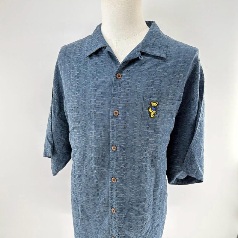 K&ouml;nnte beinhalten: Blaues kurz&auml;rmeliges Hemd mit strukturiertem Muster und holzfarbenen Kn&ouml;pfen. Eine kleine Tasche auf der Brust zeigt eine gelbe B&auml;r Grafik.