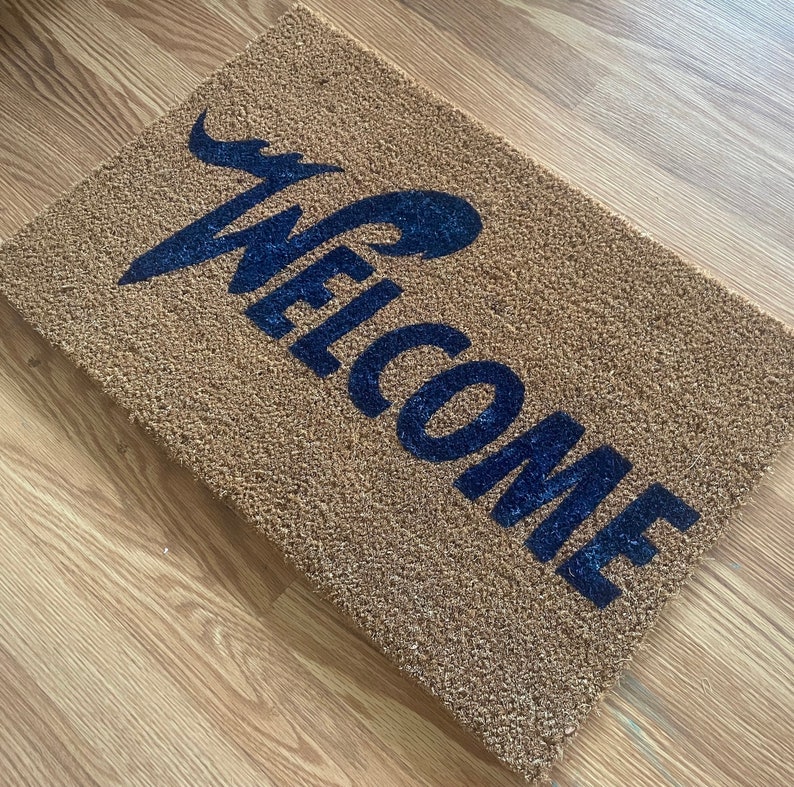 May include: A rectangular tan coir doormat featuring the word WELCOME printed in a bold, dark blue script font. The text is positioned diagonally across the coarse, natural fiber surface of the mat, creating a high-contrast visual effect.