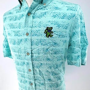 May include: A light turquoise short-sleeve button-down shirt featuring a repeating dark teal fern leaf pattern. The shirt includes a front chest pocket adorned with an embroidered green and blue dancing bear graphic and light-colored buttons.