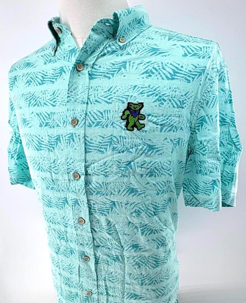 May include: A light turquoise short-sleeve button-down shirt featuring a repeating dark teal fern leaf pattern. The shirt includes a front chest pocket adorned with an embroidered green and blue dancing bear graphic and light-colored buttons.