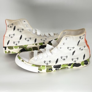 May include: Pair of high-top canvas sneakers. The white sneakers feature a camouflage pattern in green, black, and white along the soles and a bear and lightning bolt design. The back of the sneakers is orange.
