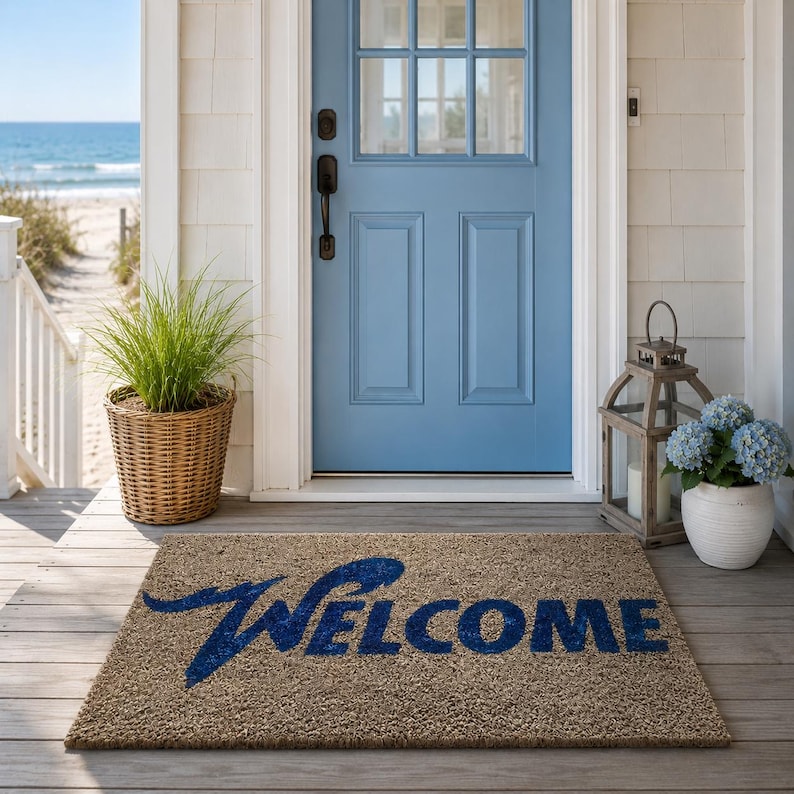 May include: A rectangular tan coir doormat featuring the word WELCOME printed in a bold, dark blue script font. The mat is positioned on a wooden porch in front of a light blue door, with a wicker basket of grass and a lantern with blue flowers.