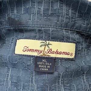 K&ouml;nnte beinhalten: Nahaufnahme eines blauen Seidenhemdes mit strukturiertem Muster. Das Hemd hat ein Etikett mit dem Text "Tommy Bahama" und "XL 100% Silk Made in China". Das Hemd ist Gr&ouml;&szlig;e XL.