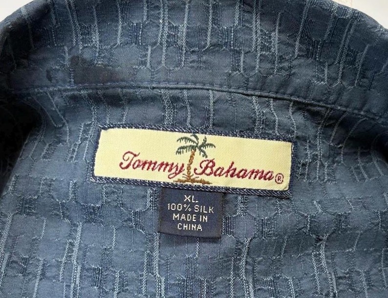 K&ouml;nnte beinhalten: Nahaufnahme eines blauen Seidenhemdes mit strukturiertem Muster. Das Hemd hat ein Etikett mit dem Text "Tommy Bahama" und "XL 100% Silk Made in China". Das Hemd ist Gr&ouml;&szlig;e XL.