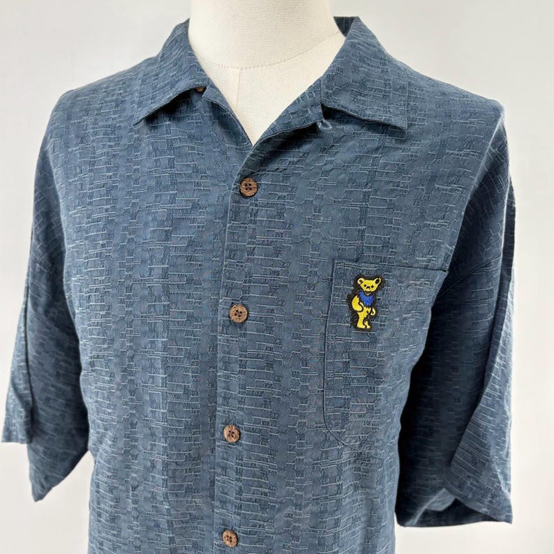 K&ouml;nnte beinhalten: Blaues kurz&auml;rmeliges Button-Down-Hemd mit strukturiertem Muster. Das Hemd hat eine Brusttasche mit einem gelben B&auml;ren-Motiv, das einen blauen Schal tr&auml;gt. Es hat Holzkn&ouml;pfe und einen klassischen Kragen.