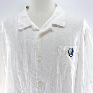 May include: White button-down shirt with short sleeves. The shirt has a chest pocket with a colorful skull and lightning bolt patch. The shirt is made of a lightweight, textured fabric and has a classic collar.