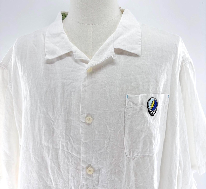 May include: White button-down shirt with short sleeves. The shirt has a chest pocket with a colorful skull and lightning bolt patch. The shirt is made of a lightweight, textured fabric and has a classic collar.