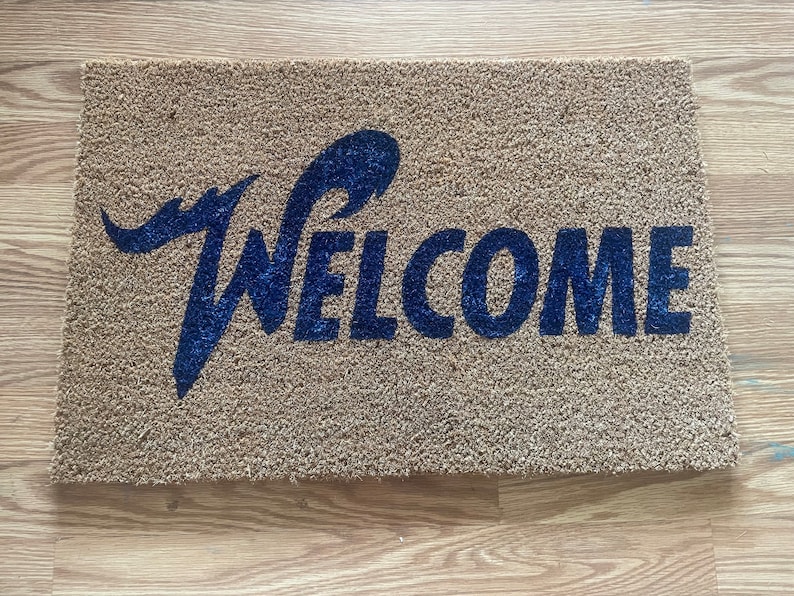 May include: A rectangular tan coir doormat featuring the word WELCOME printed in a bold, dark blue font. The letter W is stylized with an elongated, sweeping flourish that extends above the rest of the text, creating a distinct visual accent.