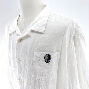 May include: White button-down shirt with a pocket featuring a colorful skull and lightning bolt patch. The shirt has a relaxed fit and a classic collar. The fabric appears to be lightweight and slightly textured.