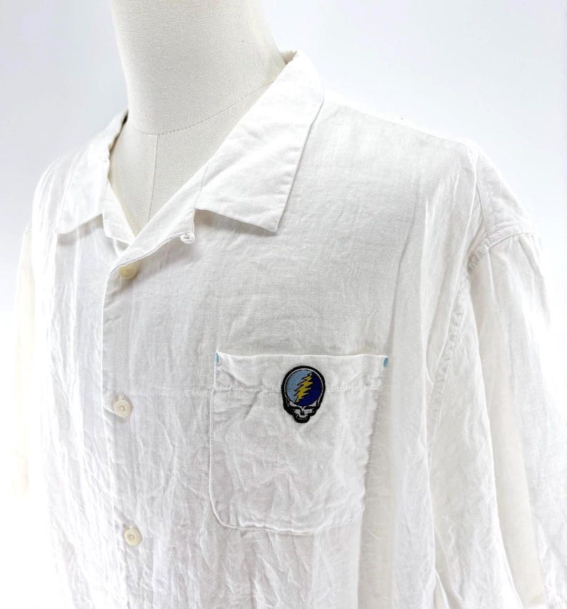 May include: White button-down shirt with a pocket featuring a colorful skull and lightning bolt patch. The shirt has a relaxed fit and a classic collar. The fabric appears to be lightweight and slightly textured.