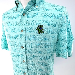 May include: A short-sleeve button-down shirt in light turquoise with a darker teal palm leaf pattern. The shirt features a button-down collar, a front chest pocket, and a small embroidered patch of a dancing bear in green, blue, and black.