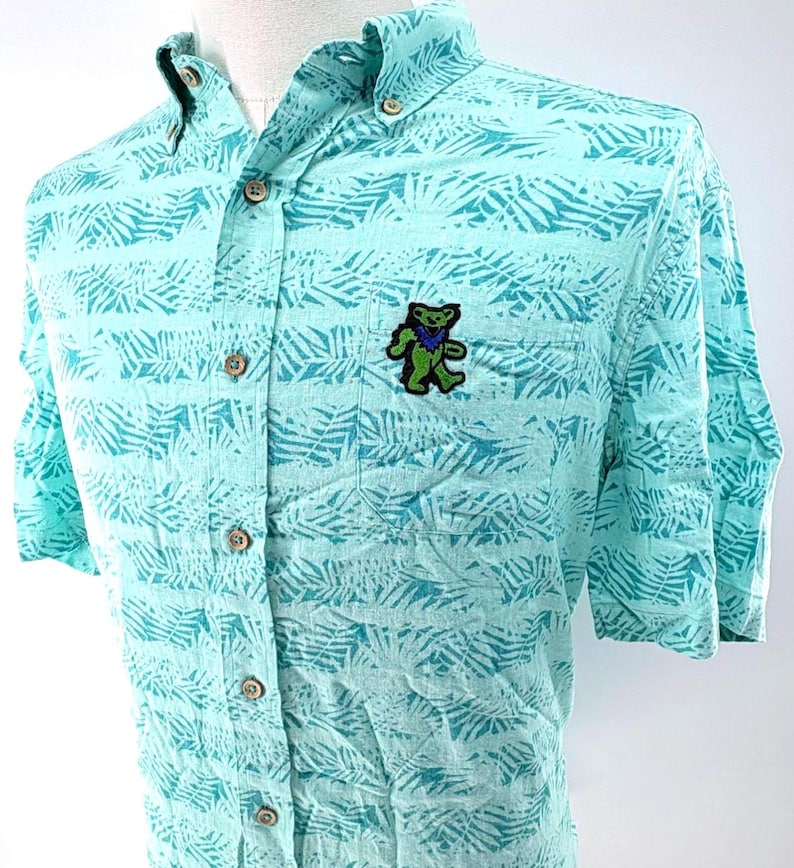 May include: A short-sleeve button-down shirt in light turquoise with a darker teal palm leaf pattern. The shirt features a button-down collar, a front chest pocket, and a small embroidered patch of a dancing bear in green, blue, and black.