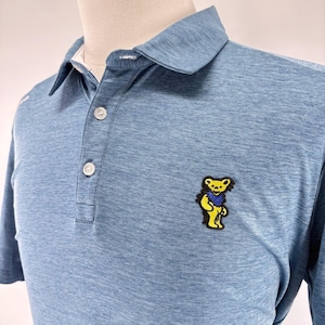 May include: Light blue polo shirt with a three-button placket and a collared neckline. The shirt features a yellow bear patch with a blue scarf on the left chest. The shirt is made of a textured fabric.
