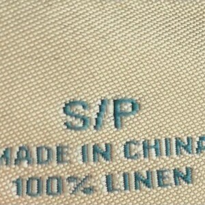 May include: Close-up of a beige fabric tag with the text "S/P", "MADE IN CHINA", and "100% LINEN" in a teal color. The tag has a textured surface.