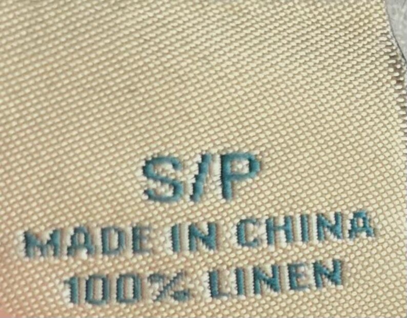May include: Close-up of a beige fabric tag with the text "S/P", "MADE IN CHINA", and "100% LINEN" in a teal color. The tag has a textured surface.
