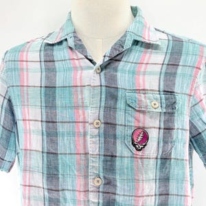 May include: A short-sleeved plaid shirt in shades of blue, pink, and white. The shirt features a button-down front, a chest pocket, and a small patch with a skull and lightning bolt design. The shirt is displayed on a mannequin.