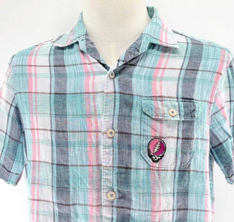May include: A short-sleeved plaid shirt in shades of blue, pink, and white. The shirt features a button-down front, a chest pocket, and a small patch with a skull and lightning bolt design. The shirt is displayed on a mannequin.