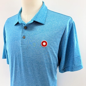 May include: Light blue polo shirt with a textured diagonal pattern. Features a small red and white circular logo on the chest and a PGA Tour logo on the sleeve. The shirt has a button-down collar and short sleeves.