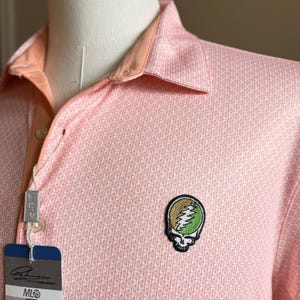 May include: A light pink polo shirt with a geometric pattern and a peach-colored collar. The shirt features a patch with a skull and lightning bolt design. A tag with the brand name is attached.