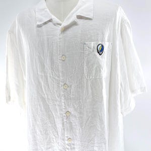 May include: White short-sleeve button-down shirt with a single chest pocket. The shirt features a colorful embroidered emblem on the pocket. The shirt has a relaxed fit and is made of a lightweight fabric.
