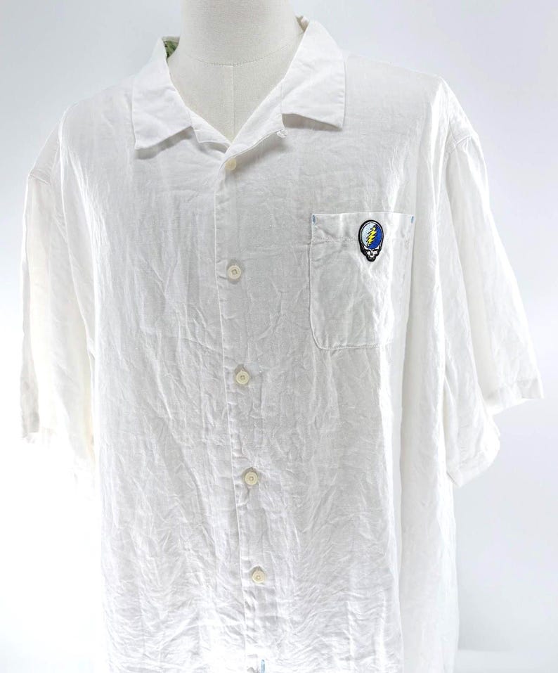 May include: White short-sleeve button-down shirt with a single chest pocket. The shirt features a colorful embroidered emblem on the pocket. The shirt has a relaxed fit and is made of a lightweight fabric.
