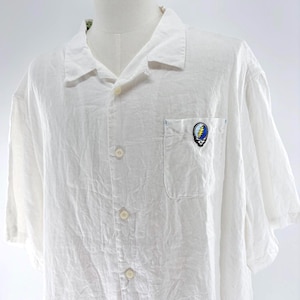 May include: White short-sleeve button-down shirt with a pocket on the chest. The pocket features a colorful emblem with a skull and lightning bolt design. The shirt is made of a lightweight fabric.