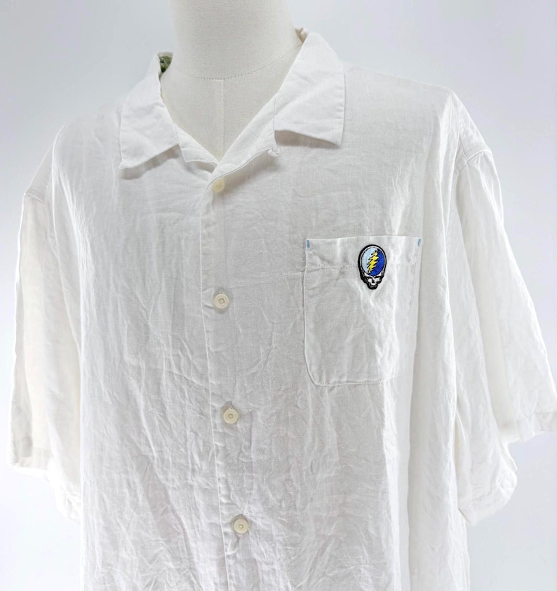 May include: White short-sleeve button-down shirt with a pocket on the chest. The pocket features a colorful emblem with a skull and lightning bolt design. The shirt is made of a lightweight fabric.