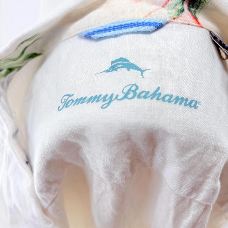 May include: Close-up of a white shirt collar with the "Tommy Bahama" logo in light blue script. The logo includes a marlin illustration. The collar has a blue and white striped detail and floral print.