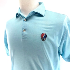 Può includere: Polo azzurro con maniche corte e colletto classico. La camicia presenta una paramontura a tre bottoni e una toppa colorata di un teschio con un fulmine e una rosa sul petto. La camicia è realizzata in un tessuto morbido e traspirante.