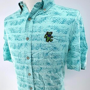 May include: A short-sleeved button-down shirt in light turquoise featuring a darker teal palm leaf pattern. The shirt includes a front chest pocket adorned with an embroidered green dancing bear patch with blue accents and a button-down collar.