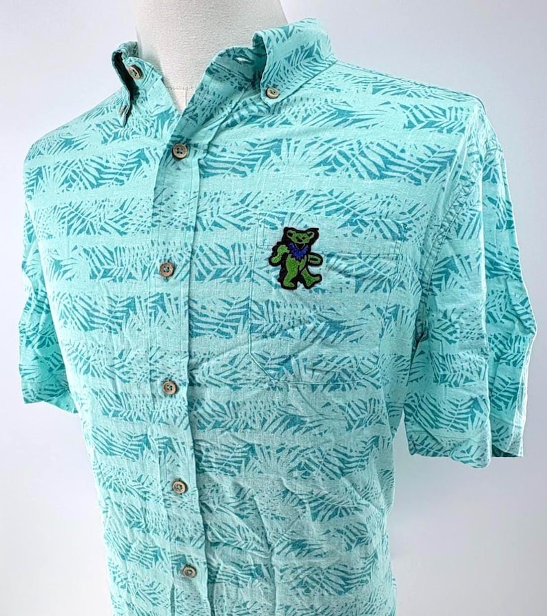 May include: A short-sleeved button-down shirt in light turquoise featuring a darker teal palm leaf pattern. The shirt includes a front chest pocket adorned with an embroidered green dancing bear patch with blue accents and a button-down collar.