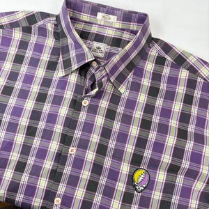 May include: A purple, black, and white plaid button-down shirt with a small embroidered patch of a smiling face with a lightning bolt on the left chest pocket. The shirt has a Peter Millar brand label on the front.