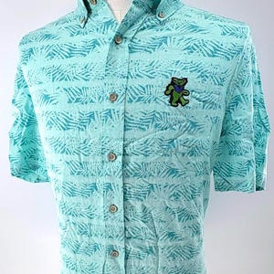 May include: A light turquoise short-sleeve button-down shirt featuring a repeating dark teal palm leaf pattern. The shirt has a button-down collar, a front chest pocket, and a small embroidered green bear graphic with a blue star on its chest.