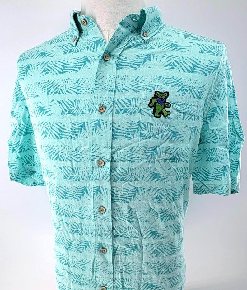 May include: A light turquoise short-sleeve button-down shirt featuring a repeating dark teal palm leaf pattern. The shirt has a button-down collar, a front chest pocket, and a small embroidered green bear graphic with a blue star on its chest.