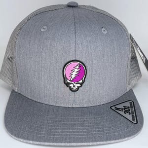 May include: Grey trucker cap with a flat peak and mesh back. It has a pink and purple skull patch on the front. The cap features a snapback closure and a tag.