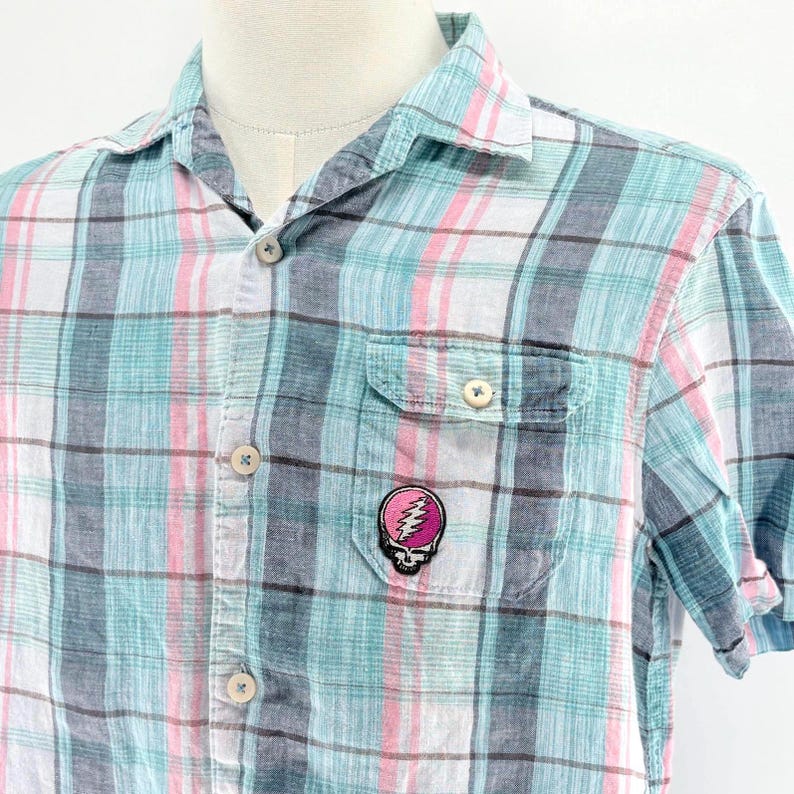 May include: Short-sleeved plaid shirt in shades of blue, pink, and white. The shirt features a button-down front, a chest pocket, and a small embroidered patch of a skull with a lightning bolt on the pocket.