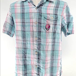 May include: A short-sleeved button-down shirt with a plaid pattern in shades of blue, pink, gray, and white. The shirt features a chest pocket with a colorful skull and lightning bolt graphic. The shirt is displayed on a mannequin.