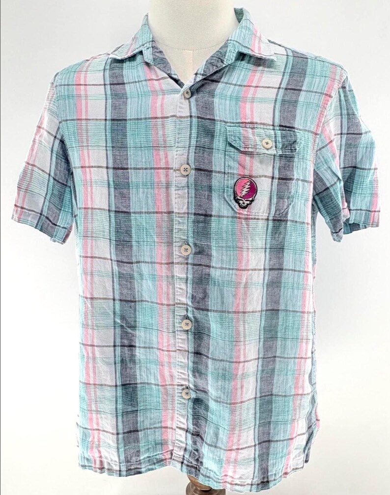 May include: A short-sleeved button-down shirt with a plaid pattern in shades of blue, pink, gray, and white. The shirt features a chest pocket with a colorful skull and lightning bolt graphic. The shirt is displayed on a mannequin.